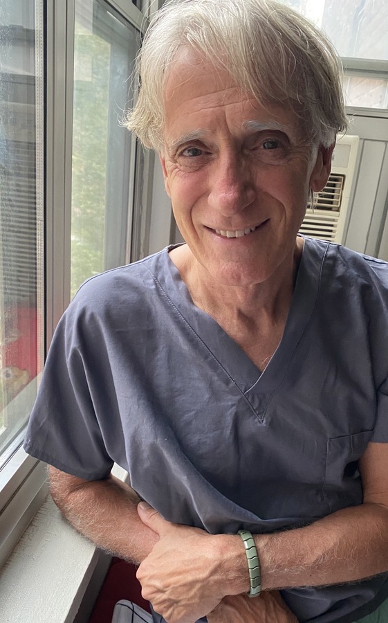 Older man with gray hair wearing gray scrubs sits by a window, smiling at the camera.