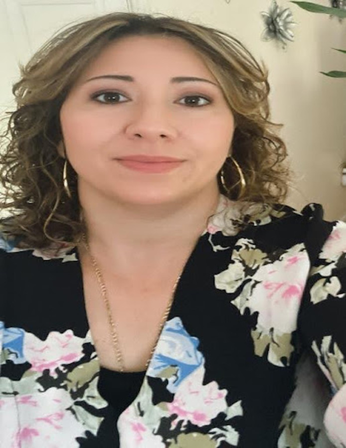 A woman with shoulder-length curly hair, wearing hoop earrings and a floral-patterned top, looks at the camera.