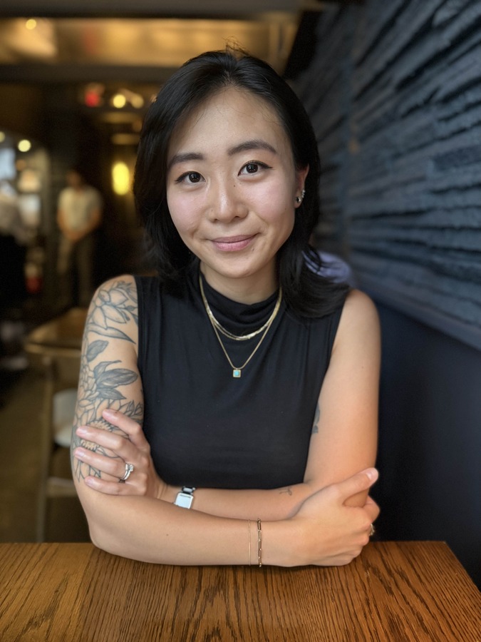 A woman with dark hair, wearing a sleeveless black top and jewelry, sits at a wooden table with her arms crossed, looking at the camera in a dimly lit restaurant.