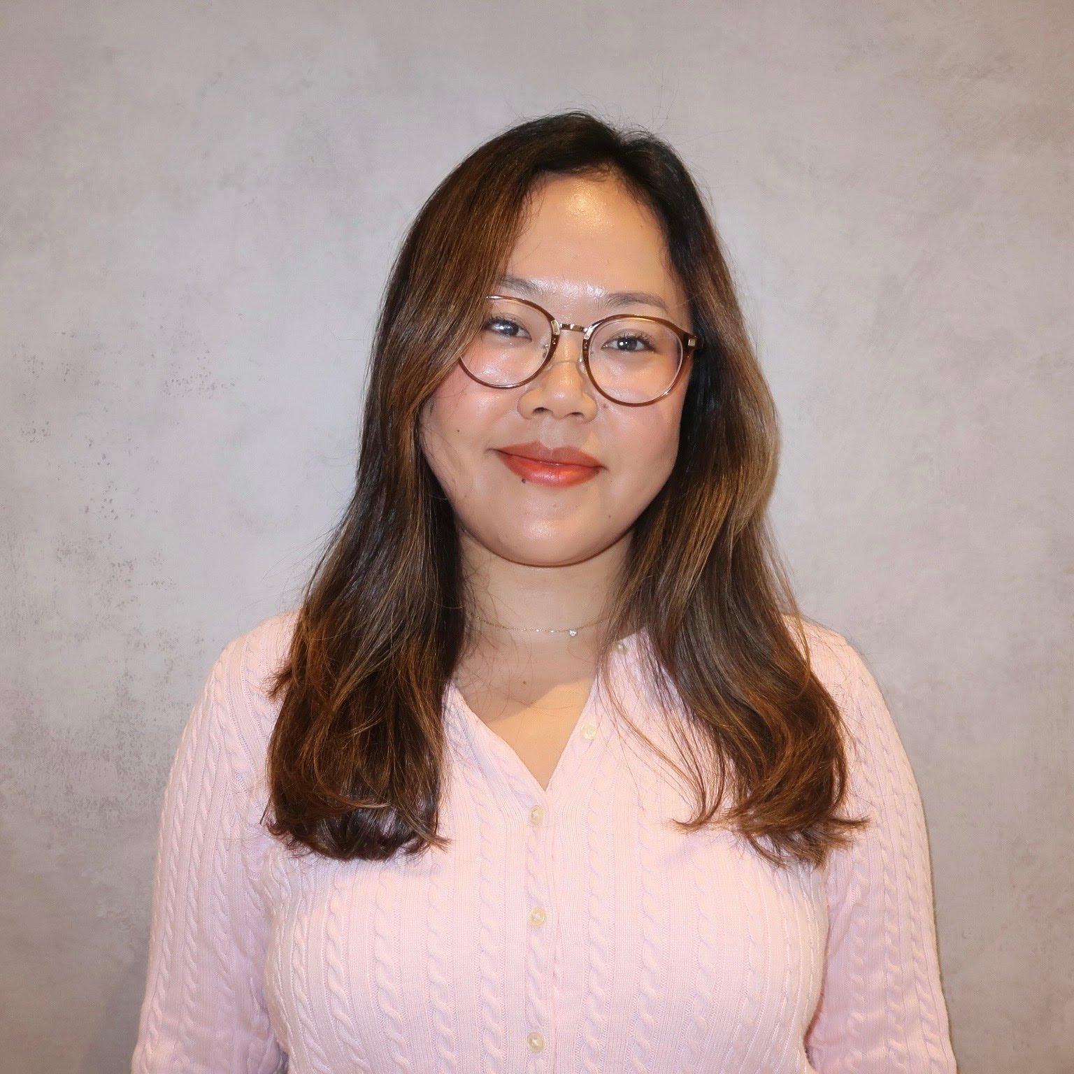 A woman with long brown hair, glasses, and a pink sweater stands in front of a plain light-colored wall, smiling slightly at the camera.
