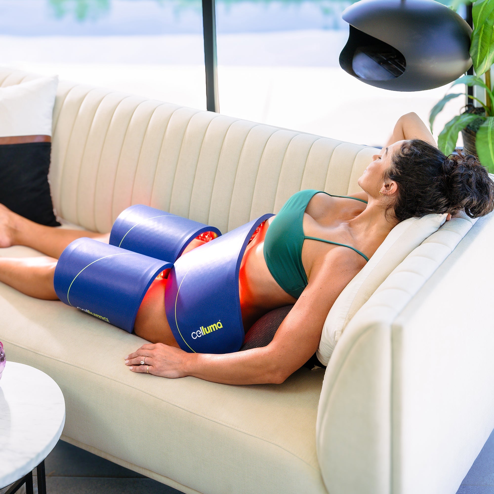 A woman lies on a sofa using Calulma light therapy devices on her midsection and thighs, wearing a green sports bra and shorts.