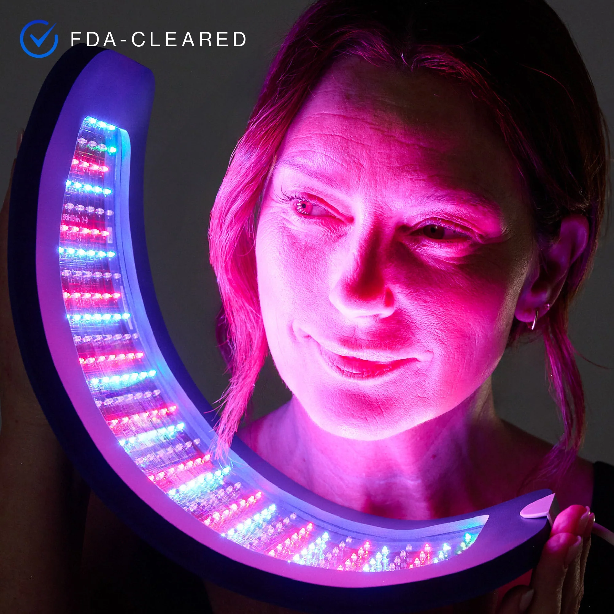 A woman holds a curved LED light therapy device emitting pink, blue, and red light near her face. A "FDA-CLEARED" label appears in the corner.