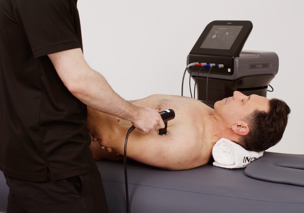 A man lies on a padded table with a towel under his head while a person uses a handheld device connected to a machine on his chest.