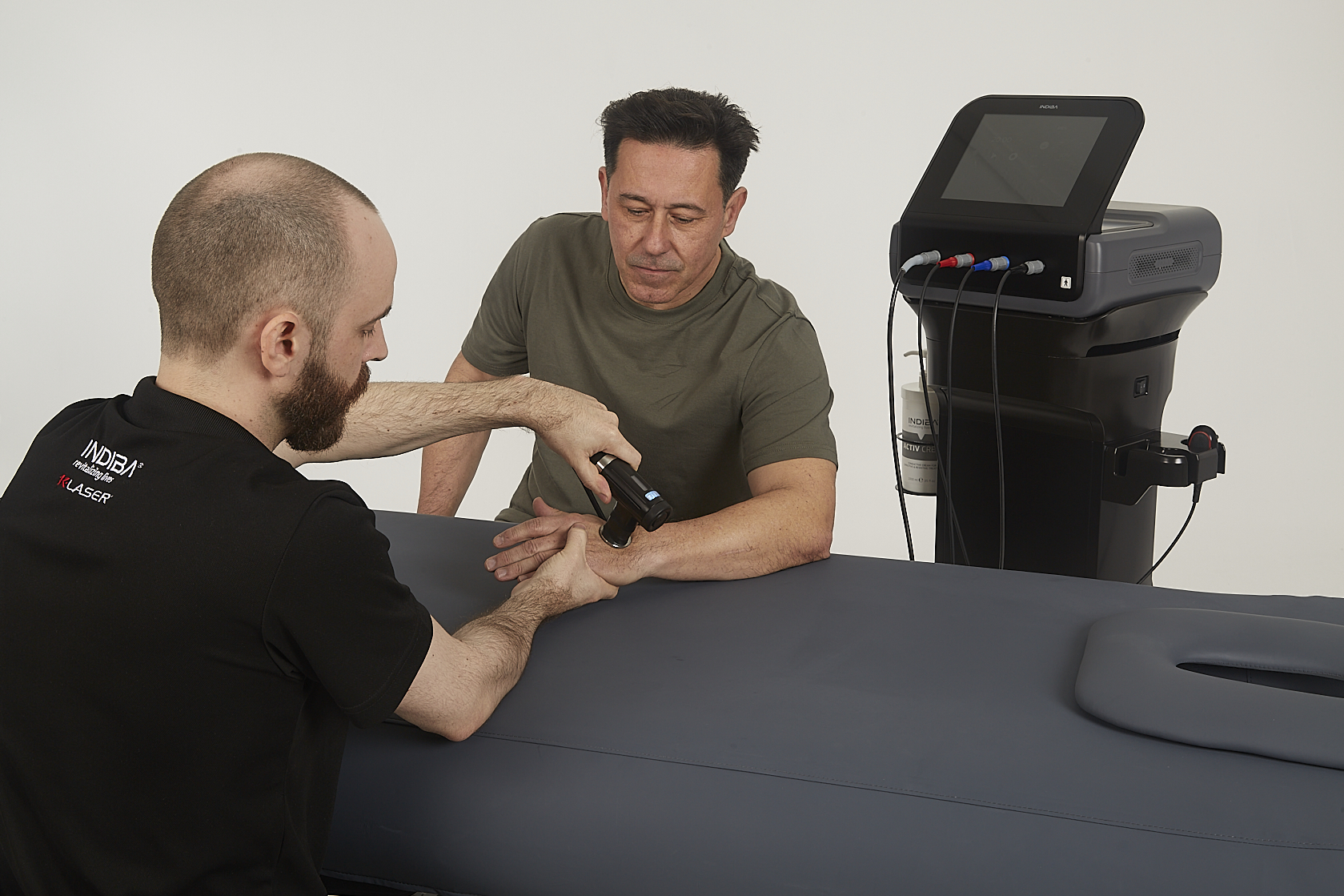A practitioner uses a handheld device on a man's hand in a clinical setting, with medical equipment visible nearby.